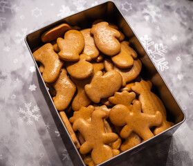 Pictured is a metal tin filled with homemade undecorated gingerbread in a variety of festive shapes such as mittens and gingerbread men. Cookies have a golden brown color and a smooth surface, placed 