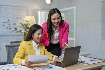Obraz premium Two happy businesswomen collaborating in the office, using a laptop while discussing their project. Analyzing financial charts and exchanging smiles, they embody teamwork and professionalism