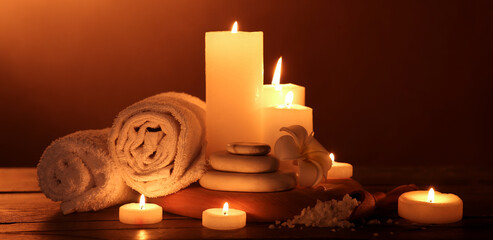 Burning candles with towels, plumeria flower, sea salt and spa stones on wooden table against dark background