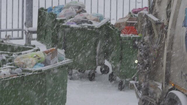 Green garbage bins in snow. Removing municipal solid waste from residential collection site with garbage truck in snowfall.