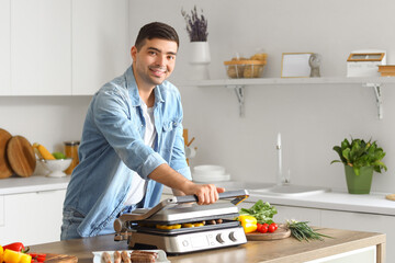 Young man cooking tasty sausages and vegetables on modern electric grill at table in kitchen