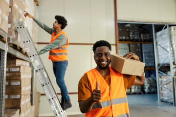 Portrait of black warehouse worker with box on shoulder giving thumbs up at camera.