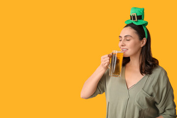 Beautiful young woman in leprechaun hat drinking beer on yellow background. St. Patrick's Day...