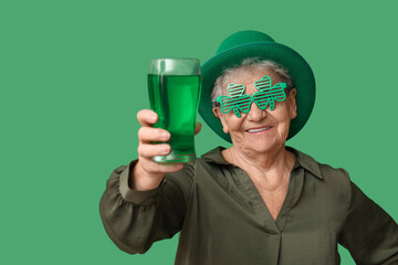 Senior woman in leprechaun hat and decorative glasses with glass of beer on green background. St....