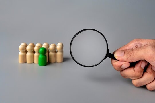 A magnifying glass is held over a row of wooden figures, with a green figure standing out from the others. Recruitment, talent acquisition concept.