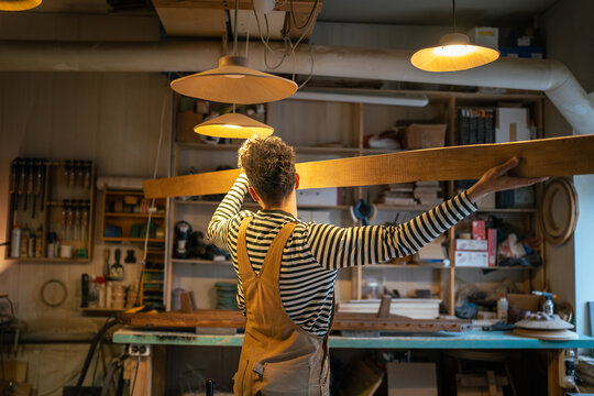 Rearview of male carpenter prepare wood beam for processing on table saw in workshop. Focused craftsmanship, precision handling of materials and attention to safe operation in furniture production