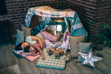 Loving mother and daughter sleeping together lying over mats in cosy diy tent. Woman embracing to little girl asleep surrounded by cushions in handmade teepee at night. Serene and peaceful atmosphere.
