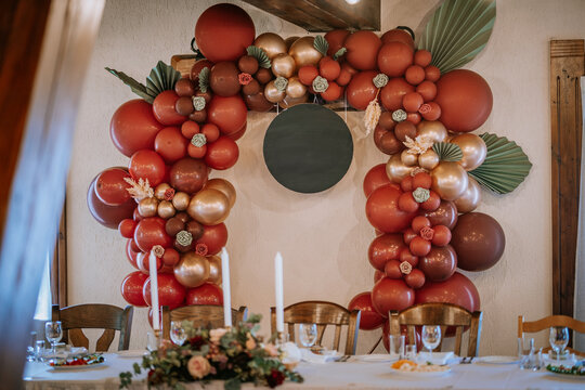 A festive balloon arch in red and gold with floral accents surrounds a blank circular sign, set above a decorated banquet table.