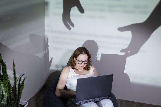 A woman intently focused on her laptop as ominous shadows loom over her, depicting the serious risks of online threats