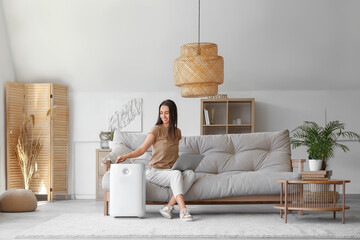 Young woman sitting on sofa and turning on modern humidifier in beautiful living room