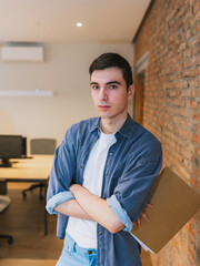 Fototapeta premium Young professional man with arms crossed, holding a folder in a modern office. Confident and determined entrepreneur ready for new challenges