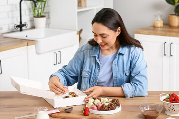 Beautiful young woman putting chocolate covered strawberries into box in kitchen