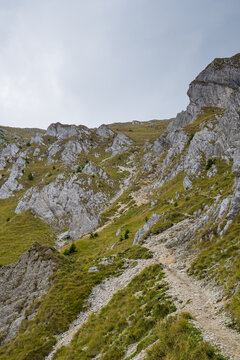 A Rugged Mountain Path Winding Through Steep Rocky Slopes And Lush Green Grass Under A Cloudy Sky