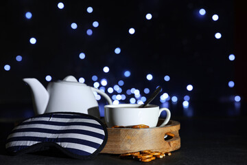 Cup and teapot of tea with pretzels and sleep mask on table against blurred lights