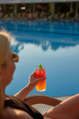 Female in sunglasses enjoys a drink on poolside. Girl chilling in tropical resort on vacation. Bright red-orange cocktail against a blue pool backdrop.