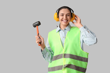 Young female construction worker with rubber mallet on grey background. Woman in male profession...
