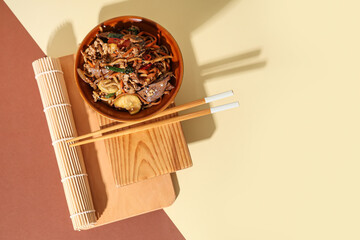 Wooden board with bowl of tasty Chinese noodles, chopsticks and bamboo mat on color background