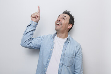 Man in casual clothes enthusiastically gestures upwards with a smile while standing against a plain...