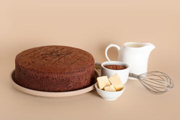 Delicious chocolate sponge cake with butter, cocoa powder, jug of milk and whisk on beige  background