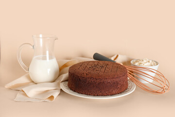 Delicious chocolate sponge cake with bowl of cream, jug of milk and whisk on beige background