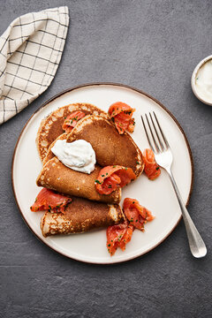 Buckwheat pancakes are garnished with fresh smoked salmon and a dollop of cream, served on a simple beige plate with a fork on the side