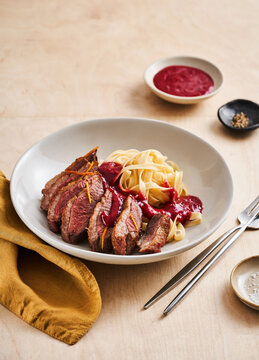 A beautifully arranged plate holds sliced beef drizzled with a deep red sauce alongside twirls of pasta. The setting is simple, with a rustic wooden table and elegant tableware
