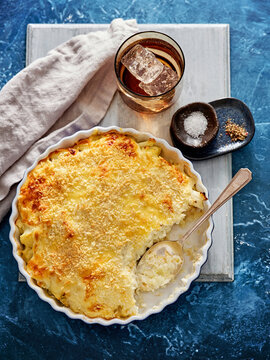 A golden brown baked vegetarian casserole occupies a white dish, accompanied by a glass with ice and two small bowls of salt and seasoning on a blue backdrop