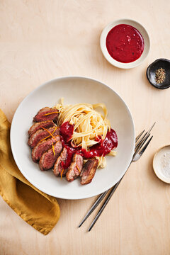 Tender steak slices are arranged next to creamy pasta on a white plate, topped with vibrant berry sauce and orange zest. Sauces in small bowls complement the dish