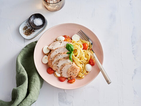 Enjoy a flavorful pasta dish featuring grilled chicken slices, fresh tomatoes, and mozzarella balls, presented in a soft pink bowl. Perfect for a relaxed meal
