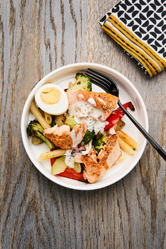 Colorful bowl filled with fresh vegetables, pasta, and grilled salmon, topped with a creamy dressing. A hard-boiled egg adds extra protein to the meal