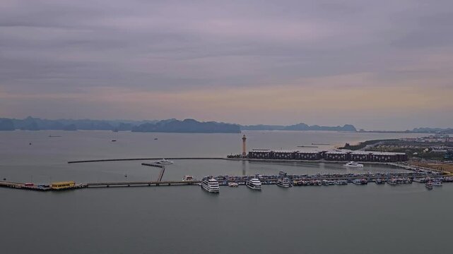 A serene aerial perspective captures Ha Long's bustling marina with its iconic white lighthouse, scattered vessels in the calm waters, and towering skyscrapers nestled against hazy karst formations