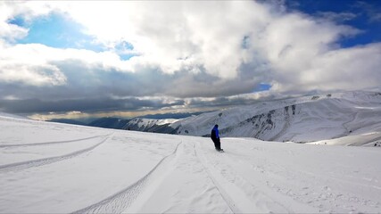 Following a snowboarder in black and blue outfit riding down the slope. Sportsman approaches a group of people standing and falls near them. Spectacular mountains and dramatic cloudscape at backdrop.