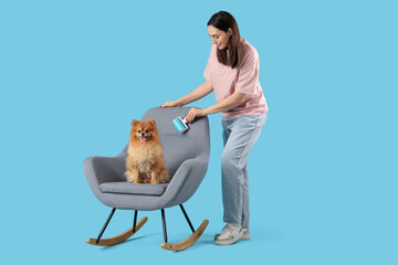 Young woman with lint roller and fluffy dog in rocking chair on blue background