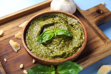 Wooden board with bowl of delicious pesto sauce and ingredients on blue background