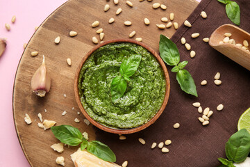 Wooden board with bowl of delicious pesto sauce and ingredients on pink background
