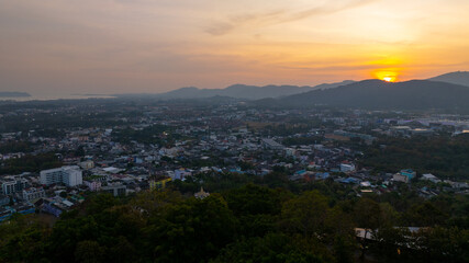 A scenic view of a town nestled among lush green mountains at sunset, with a temple structure perched on the hillside and a vibrant orange sky creating a warm, tranquil atmosphere.