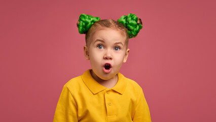 Little girl with astonished expression, mouth open in surprise, wearing yellow shirt and green braided buns against pink studio background. Concept of childhood, emotions, lifestyle