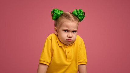 Little girl with a pouty face, arms crossed, wearing yellow shirt and green braided buns against pink studio background. Dissatisfaction. Concept of childhood, emotions, lifestyle