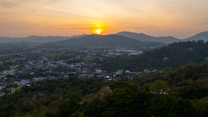 A scenic view of a town nestled among lush green mountains at sunset, with a temple structure perched on the hillside and a vibrant orange sky creating a warm, tranquil atmosphere.