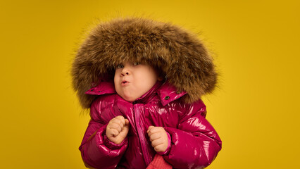 Girl wearing shiny pink coat with large fur hood, making playful expressions, against yellow studio background. Cold winter weather. Concept of childhood, emotions, lifestyle