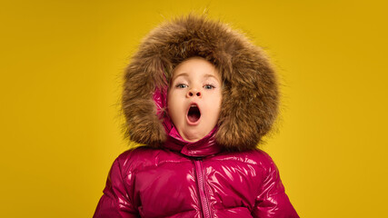 Portrait of little girl, child in pink warm winter coat yawning, feeling sleepy against yellow studio background. Concept of childhood, emotions, lifestyle