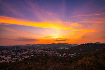 Obraz premium A scenic view of a town nestled among lush green mountains at sunset, with a temple structure perched on the hillside and a vibrant orange sky creating a warm, tranquil atmosphere.