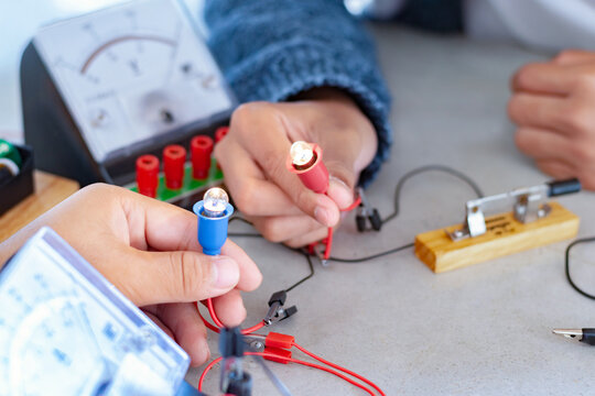 Children are studying electrical circuit systems by experimenting with electrical connections using a small electric meter and a small flashlight bulb in physics science class, soft focus.