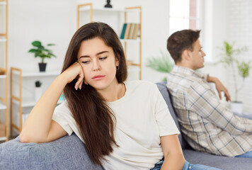 Upset family couple sitting on sofa in living room back to back and ignoring each other. Frustrated young woman sitting separately and man in background facing away from her after quarrel.