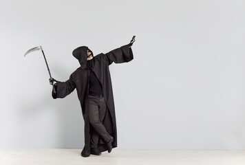 Happy skeleton in a grim reaper death costume holding scythe, doing funny pose isolated on studio...
