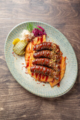 A beautifully plated dish featuring grilled sausages drizzled with sauce, served on a wooden surface. Accompanied by pickles, cabbage, and garnishes for a colorful presentation.