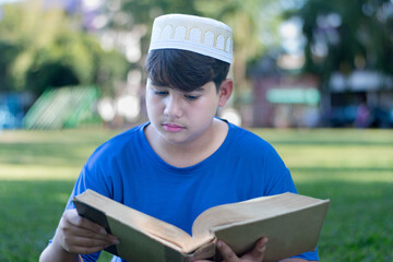 Asian Muslim boy is reading al-quran book in the green grass lawn in the evening, soft and...