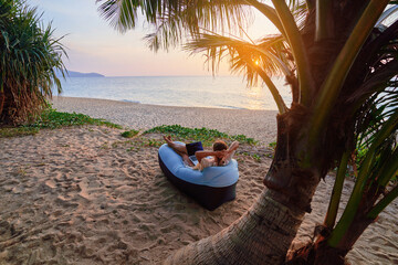 Young contented happy girl digital nomad freelancer lies on an inflatable sofa under a palm tree on...