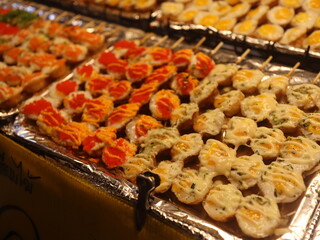 A delicious assortment of skewered snacks displayed on a shiny foil tray. Topped with colorful ingredients like cheese, herbs, and sauces, the scene captures the essence of a vibrant food market.