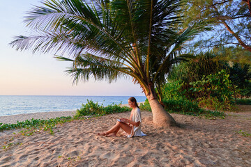 Young contented joyful happy girl digital nomad freelancer sitting on a sand under a palm tree on...